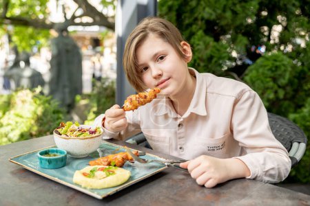 Photo for Caucasian child in cafe eats chicken skewers with appetite. Teen boy has breakfast in restaurant. Kid holding kebab - Royalty Free Image