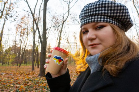 Photo for Ukraine. Autumn evening. Sunset. City Park. End of the working day. The girl holds in her hand a cup of coffee with a patriotic sticker "I love Ukraine". Walks in the open air - Royalty Free Image