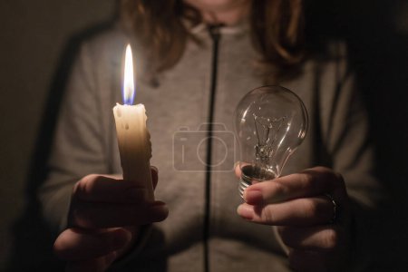 Stromausfall. Zerstörung der Infrastruktur. Energiekrise. Stromausfall-Konzept. Das Mädchen hält eine Glühbirne und eine brennende Kerze in ihren Händen. Nahaufnahme
