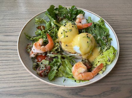Foto de Desayuno LCHF saludable. Huevos Benedicto con quinua, ensalada fresca, camarones y salsa en un plato beige sobre una mesa de madera - Imagen libre de derechos