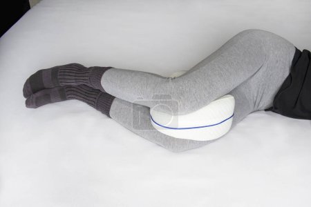 Téléchargez les photos : Young woman in pajama pants with an anatomical pillow between her legs and knees, lying on a bed with white sheets - en image libre de droit