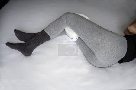 Foto de Young woman in pajama pants with an anatomical pillow between her legs and knees, lying on a bed with white sheets - Imagen libre de derechos