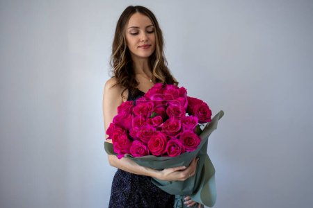 A beautiful young woman in festive clothes with curly hair holds a large bouquet of roses and looks on. Slender girl.