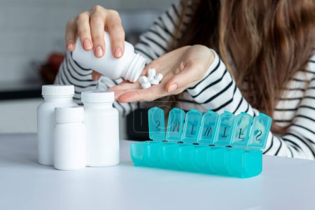 Foto de A young woman pours white pills into her hand in front of a pill organizer. Nearby mock-ups of white cans of supplements - Imagen libre de derechos