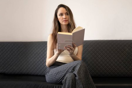 Die schöne, grübelnde junge Frau sitzt auf der Couch, hält ein aufgeschlagenes Buch in der Hand und schaut weg..