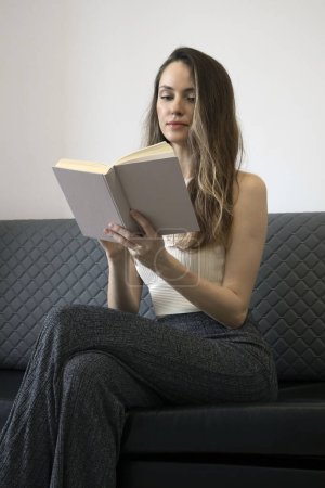 Téléchargez les photos : Femme en blanc et gris assis sur un canapé et lit un livre de papier. Femmes en littérature. - en image libre de droit
