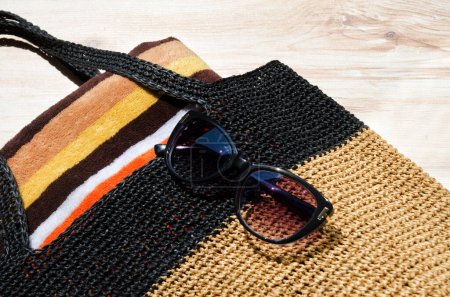 Schwarze Strandtasche aus Bast. Frauentasche mit Handtuch und Sonnenbrille auf dem Tisch.