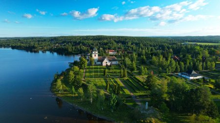 Aerial view of 15th century church and cemetery. Historical monument by the lakeside of Siljan lake. Mountains and forests in the background, during sunrise golden hour, in the Swedish countryside