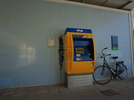 Foto de Máquina expendedora de billetes en la estación de tren de Nederlandse Spoorwegen NS (Ferrocarriles Holandeses). En los colores azul amarillo con el logotipo de NS. Y una bicicleta holandesa tradicional. Amsterdam, Países Bajos, 05-04-2023 - Imagen libre de derechos