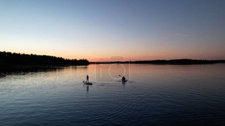 Photo for Silhouette of mother with kids sitting and standing on inflatable sup boards, paddling on calm lake bysjon during blue hour. Active water sport summer vacation. Nas bruk Dalarna Sweden. Aerial view - Royalty Free Image
