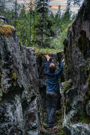 Téléchargez les photos : Young Explorer Scaling a Forest Crevasse in Sweden. Un garçon grimpant soigneusement entre les murs rocheux imposants recouverts de mousse, entouré par la végétation dense d'une forêt suédoise sous le ciel du soir d'été. - en image libre de droit