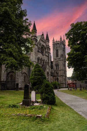 Majestic Nidaros Cathedral Tower Facade by Green Field and graveyard. A grand church with a tall tower stands next to a lush green field on a beautiful summer evening. In Trondheim Norway