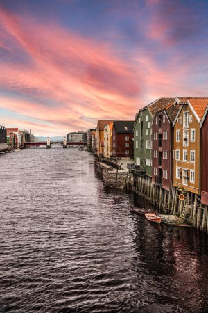 Ein ruhiger Abendhimmel mit rosa und orangen Farbtönen über den bunten historischen Gebäuden auf Stelzen am Ufer des Flusses Nidelva. Touristisches Reiseziel in Trondheim, Norwegen.
