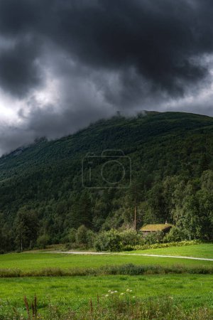 Un campo verde con cabaña de bosque nórdico al pie de una montaña boscosa y nubes de tormenta, mostrando la belleza de la naturaleza noruega en Más og Condado de Romsdal