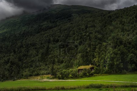 Secluded Norwegian Hut at Mountain Base Before a Storm. A small wooden summer cabin nestled in a forest clearing, with dark storm clouds gathering over a mountain peak. In the Nordic wilderness of Norway