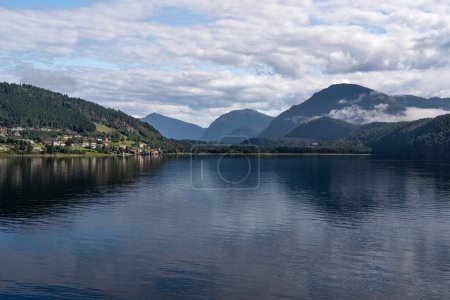 Calm waters of Stangvikfjorden mirror the sky, with the picturesque village of Kvanne nestled against a mountain, overlooking the Trollheimen mountains. Scandinavian travel destination in summer