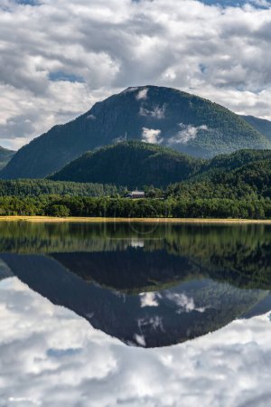 Reflections on the calm flat water at the Stangvikfjord with a view of the Soysetoran nature reserve and a farm in the mountains of Trollheimen, More og Romsdal Norway. A serene fjord reflects the lush green mountains and white clouds on a summer day