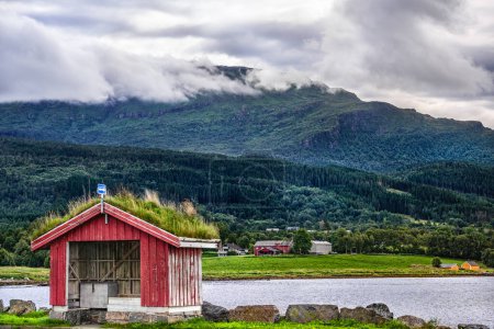 Un abri de bus rouge pittoresque avec un toit de gazon vert luxuriant se dresse près de Halsafjord dans un contexte de montagnes boisées et de nuages spectaculaires en été, à Halsa en Norvège