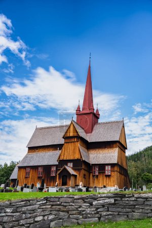 Blick auf die mittelalterliche Ringebu Stabkirche aus dem 13. Jahrhundert steht inmitten von Grabsteinen unter blauem Himmel mit Wolken im Sommer, die Jahrhunderte des norwegischen Erbes widerspiegeln.