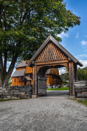 A wooden gate leading to the historic medieval Ringebu Stave Church surrounded by headstones against a summer blue sky with clouds Innlandet county, Norway