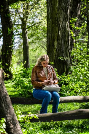 Attraktive Frau, die in einem Park ferngesteuert mit ihrem Handy arbeitet. Eine Frau sitzt auf einer Parkbank und verschickt eine E-Mail von ihrem Smartphone
