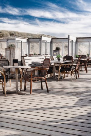 Serene beach pavilion terrace overlooking dunes on a soft blue cloud sky day. A vacant terrace of a beachside pavilion, with chairs and tables set against a backdrop of sand dunes.