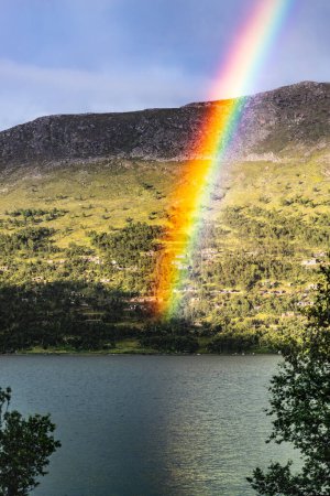 Un arco iris radiante arcos sobre un sereno lago noruego, aterrizando cerca de las cabañas de troncos tradicionales enclavadas entre las colinas de Oppdal, Noruega
