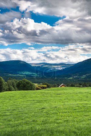 Saftiges Grasland im Oppdal-Tal in Trondelag Norwegen mit einem Bauernhof versteckt hinter einem sanften Hügel, unter einem Himmel voller Wolken.