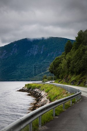 A solitary car journeys on Norwegian National Road Highway 70 beside Alvundfjord, bordered by lush greenery and a mountain in the distance under a cloudy sky.