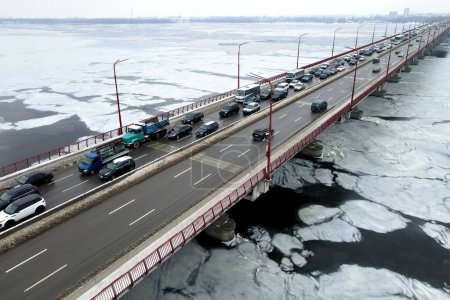 Photo for Many cars drive over river bridge in winter. Traffic jam at rush hour, air pollution, smog on highway, road. New bridge in Dnipro city, Ukraine, aerial drone view - Royalty Free Image