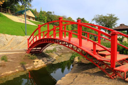 Old red wooden bridge across small river in park. Vintage Japanese style bridge, water. Uman park, Ukraine in summer, spring, autumn.