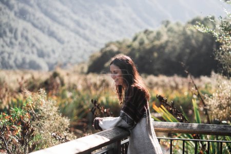 Téléchargez les photos : Fille brune caucasienne en chandail de laine marron et gris calme et détendu en regardant le paysage vert des montagnes et des arbres depuis un balcon en bois, Lake Gunn, Nouvelle-Zélande - Concept de voyage - en image libre de droit