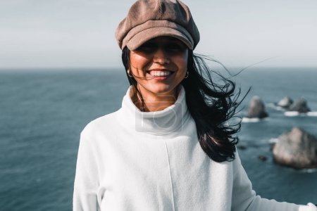 Foto de Joven mujer caucásica en suéter blanco y bonitos pendientes en sus oídos sonriendo a la cámara desde el mirador cerca de las olas del océano, punto de pepita, Nueva Zelanda - Concepto de viaje - Imagen libre de derechos
