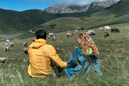 Téléchargez les photos : Caucasien garçon et fille assis ensemble sur l'herbe du champ en regardant les vaches serrant calmement la main par une journée ensoleillée dans la nature, ruta del cares asturias, espagne - Lieux traditionnels espagnols - en image libre de droit