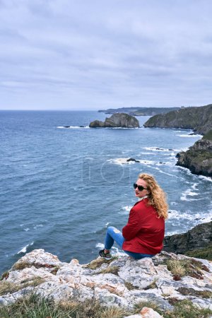 Foto de Mujer joven rubia caucásica con chaqueta roja y pantalones azules zapatillas gafas de sol sentadas en una roca mirando a la cámara junto a los acantilados y las olas del mar, cabo de penas, España - Paisajes españoles - Imagen libre de derechos