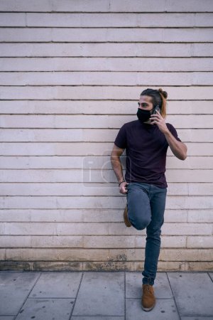 Foto de Caucásico chico de pie en negro camiseta marrón zapatos apoyados en la pared sucia en la ciudad hablando en el teléfono inteligente con máscara facial - Estilo urbano - Imagen libre de derechos