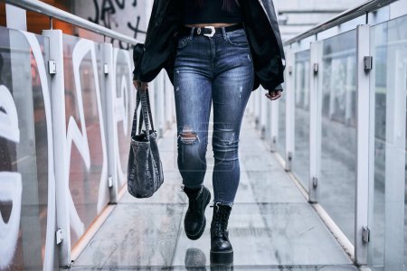 Foto de Frente a las piernas de una chica latina en pantalones azules rasgados chaqueta de piel y grandes botas negras caminando por la pasarela de cristal - Concepto urbano - Imagen libre de derechos