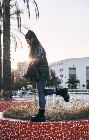 Photo for Young latin brunette woman with long ponytail in her hair blue pants standing on a small garden and big boots next to palm trees with one foot in the air in urban park - Urban concept - Royalty Free Image