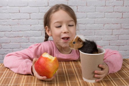 Photo for Cheerful child girl with a bitten apple in her hand and a guinea pig sticking out of a cardboard glass, close-up - Royalty Free Image