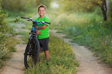 Foto de Happy boy stands in nature and smiles, holds a bike, is going to ride, beautiful sunlight and bokeh - Imagen libre de derechos