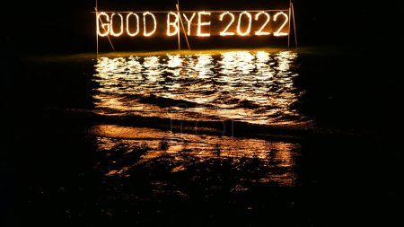 Téléchargez les photos : Adieu 2022 phrase brûlante faite de bâton de feu placé sur l'eau dans la mer, joyeuse célébration de la nouvelle année dans un lieu tropical la nuit, inscription sur la plage - en image libre de droit