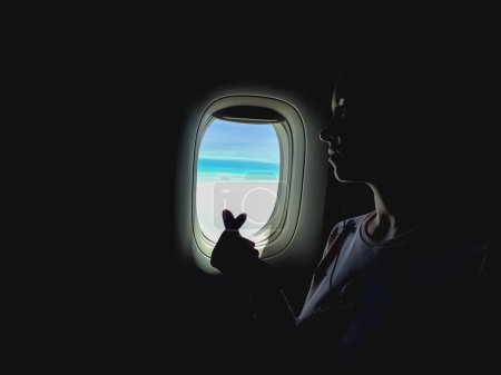 Photo for Creative photo in dark tones, woman looking out of airplane window, flying on aircraft. Female crossed fingers like korean heart sign. Love to travel - Royalty Free Image