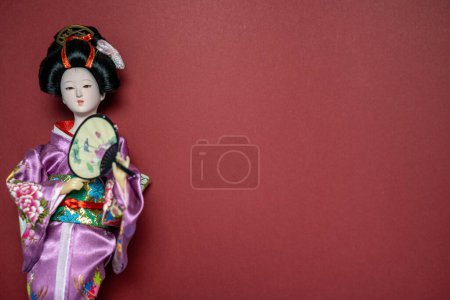 Téléchargez les photos : Asian ceramic doll in traditional clothes, holds fan in hands, red background, copy space, place for text, oriental culture - en image libre de droit