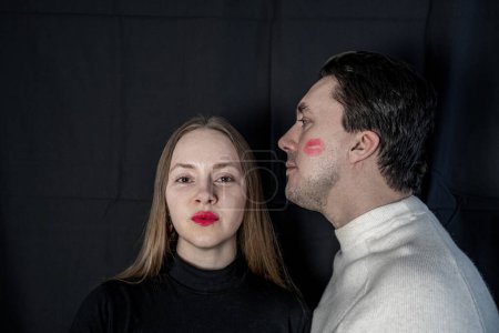Photo for Couple on dark background, woman in black, no make up, natural look with red lips, looking at camera. Man in white, looks at wife. Lipstick print on cheek. Family, yin yang contrast, opposites attract - Royalty Free Image