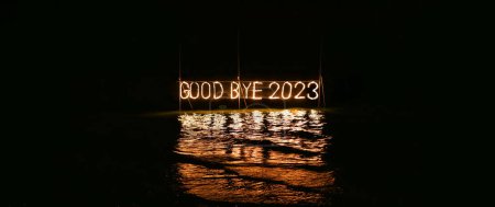 Good bye 2023 illuminated sign installation in water on the beach on black background. Old year is burning. Happy winter holidays made of lights and garland. Street advertisement. Outdoor signboard. 