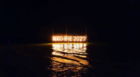 Téléchargez les photos : Au revoir 2023 installation de panneaux lumineux dans l'eau sur la plage sur fond noir. La vieille année brûle. Joyeuses vacances d'hiver faites de lumières et guirlande. Publicité de rue. Panneau extérieur. - en image libre de droit