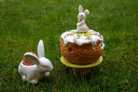Photo for White Easter rabbit standing on home-baked easter cakes with icing on a plate on green background. Religious holiday. Rabbit with dyed egg - Royalty Free Image