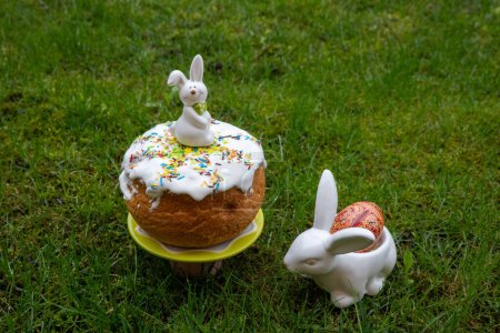 white Easter rabbit standing on home-baked easter cakes with icing on a plate on green background. Religious holiday. Rabbit with dyed egg