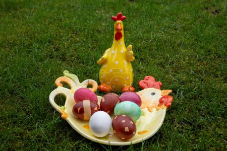 Photo for Ceramic rooster near dyed easter eggs on the chicken-shaped plate on green background. Easter celebration. - Royalty Free Image