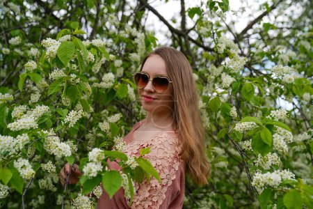 hermosa rubia mujer de pelo natural lleva gafas de sol en traje de color rosa está posando en el parque botánico jardín cerca de flor de cerezo árbol de mayday con flores. Primavera y pureza, belleza natural